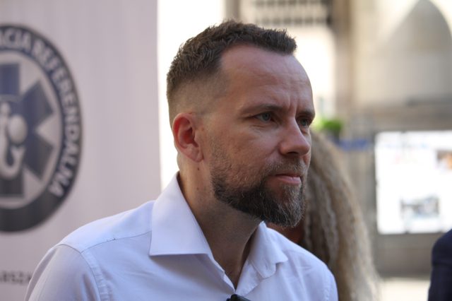 Maciej Wilk w białej koszuli na tle bannera Stołecznej Operacji Referendalnej