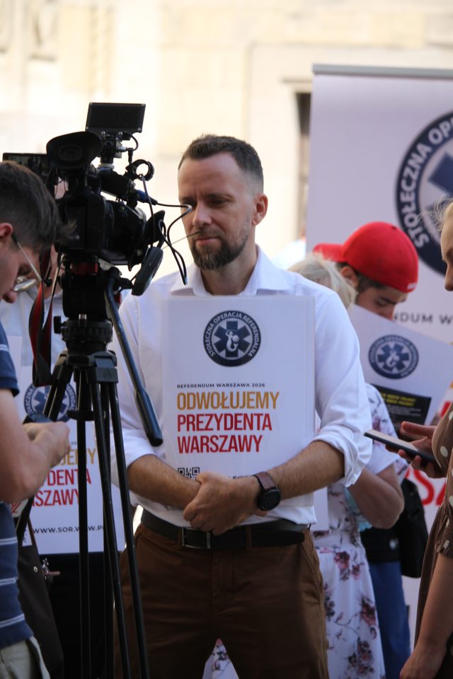 Maciej Wilk z plakatem Stołecznej Operacji Referendalnej otoczony dziennikarzami