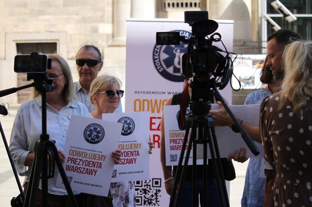 Wolontariusze z plakatami "Odwołujemy prezydenta Warszawy" przed kamerami mediów